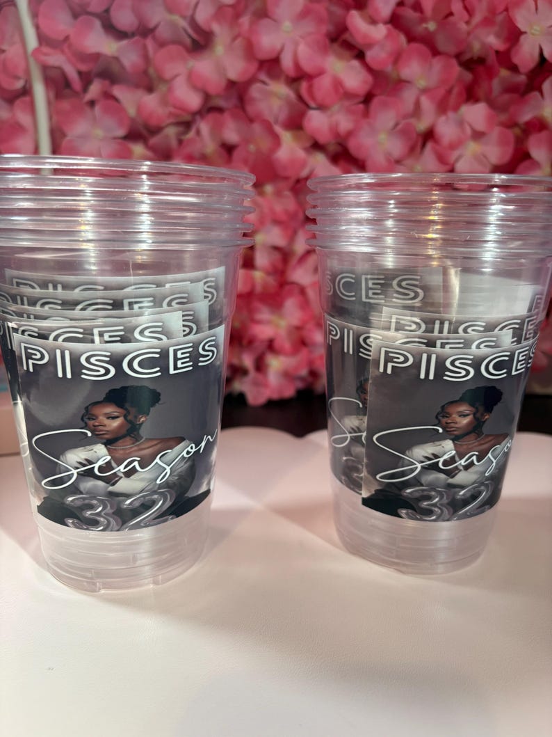 May include: Two clear plastic cups with a printed design. The design features the word "PISCES" and "Season 32" with an illustration of a woman. The background is a blurred display of pink flowers.
