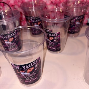 May include: Clear plastic cups with a printed label that reads "N-Valley Nelly's Bash" in white and pink. The cups are arranged on a white surface, with a backdrop of pink flowers. The cups are empty.