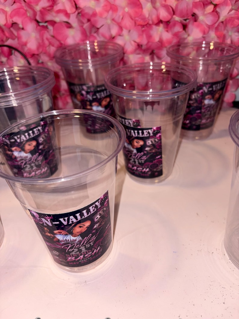 May include: Clear plastic cups with a printed label that reads "N-Valley Nelly's Bash" in white and pink. The cups are arranged on a white surface, with a backdrop of pink flowers. The cups are empty.