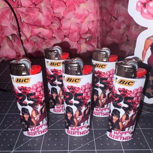 May include: Five white BIC lighters featuring a custom wrap design with pink balloons, the number 25 in black, a photograph of a person, and the text HAPPY BIRTHDAY. Each lighter has a metallic top and a red ignition button for standard use.