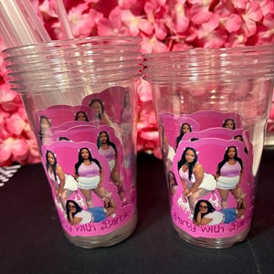 May include: Two clear plastic cups with pink and white labels featuring a woman in various poses. The labels read "Party with Barbie". Clear straws are visible in the left cup. The background is a pink floral arrangement.