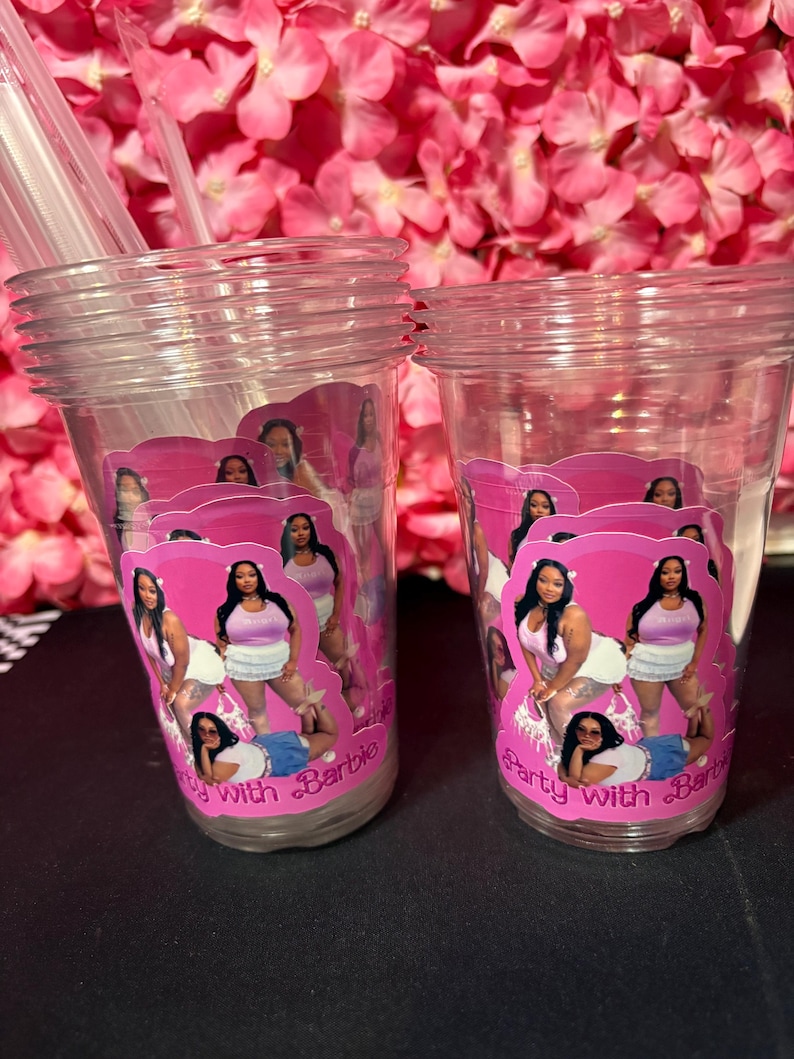 May include: Two clear plastic cups with pink and white labels featuring a woman in various poses. The labels read "Party with Barbie". Clear straws are visible in the left cup. The background is a pink floral arrangement.