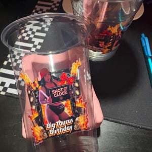 May include: Clear plastic cups with a colorful graphic. The graphic features a person holding a sign that says "Shot O' Clock" and "Big Taurus Birthday" with flames and skulls.