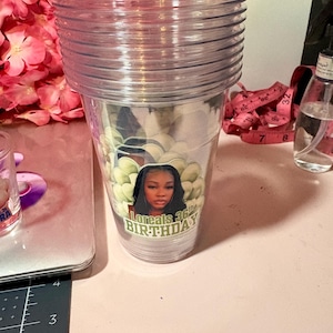 May include: A stack of clear plastic tumblers. The bottom tumbler has a sticker with a woman's image and the text "Loreals 36th BIRTHDAY". The tumblers are on a pink surface with pink flowers in the background.