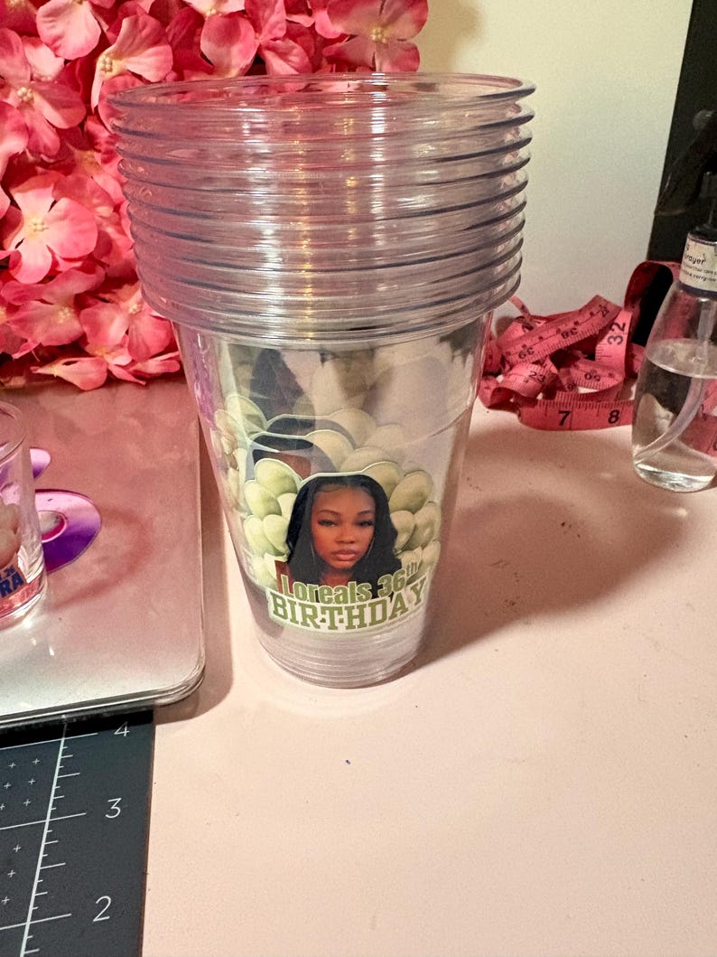 May include: A stack of clear plastic tumblers. The bottom tumbler has a sticker with a woman's image and the text "Loreals 36th BIRTHDAY". The tumblers are on a pink surface with pink flowers in the background.
