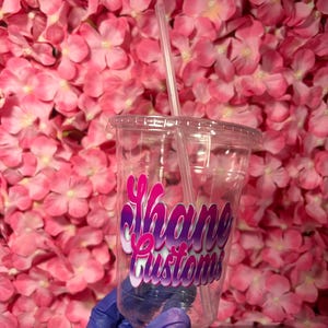May include: A clear plastic cup with a lid and straw, featuring the text "Shane Customs" in a gradient pink and purple design. The cup is held against a backdrop of pink flowers.