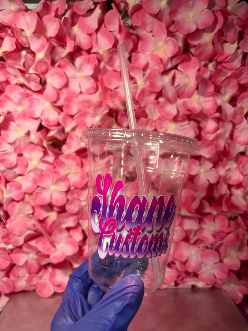 May include: A clear plastic cup with a lid and straw, featuring the text "Shane Customs" in a gradient pink and purple design. The cup is held against a backdrop of pink flowers.