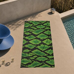May include: A rectangular beach towel with a vibrant green and black snakeskin pattern. The towel is lying on a concrete surface near a pool. The towel's design features a repeating diamond-shaped pattern.