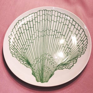 May include: A round white ceramic dinner plate featuring a large, centered graphic illustration of a scallop shell rendered in a deep forest green line art style. The design covers the majority of the plate surface with intricate details.