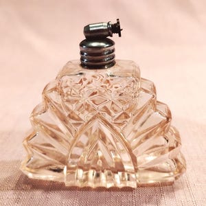 May include: A clear glass perfume bottle with a stepped, fan-like geometric shape and a textured diamond pattern on the front. The bottle features a silver-toned metal screw-top cap with a small, protruding cylindrical nozzle at the very top.