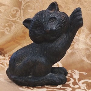 Può includere: Statuetta di gatto in ghisa nera in posizione seduta, con una zampa alzata. Il gatto presenta dettagli precisi e una finitura leggermente usurata, che gli conferisce un aspetto vintage. L'oggetto è decorativo.