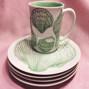 May include: A white ceramic mug and a stack of three white ceramic saucers, all decorated with green line-art illustrations of various seashells. The mug features a solid light green interior and a handle on the right side, while the top saucer displays a large, detailed fan-shaped shell pattern.