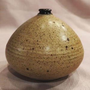 May include: A small, round ceramic vase with a speckled tan and beige glaze. The body of the vase features subtle horizontal ridges along the sides, and it has a narrow, dark brown rim at the top opening, creating a distinct contrast with the lighter base.