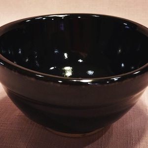 May include: A deep, round ceramic bowl with a glossy, solid black glaze. The bowl features a smooth, curved interior and a slightly tapered exterior wall that leads down to a small, unglazed circular base showing a light tan clay color.