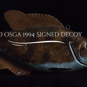 May include: A brown and green fish decoy with the text "FLOYD OSGA 1994 SIGNED DECOY". The fish has detailed fins and a slightly open mouth. It is set against a black background.