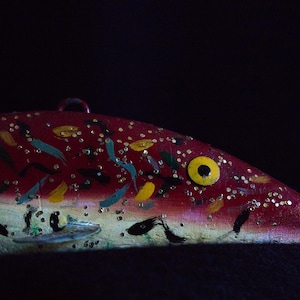 May include: A hand-painted wooden fish ornament with a red body, decorated with yellow, blue, and gold accents. The fish has a large yellow eye and a small loop for hanging. The background is black.