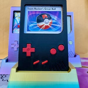 Op de afbeelding: Een zwarte Game Boy-vormige kaarthouder met rode knoppen en een D-pad, met een "Team Rocket's Great Ball"-kaart. De houder staat op een veelkleurige basis, met twee vergelijkbare houders op de achtergrond.