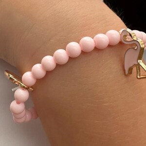 May include: A pink beaded bracelet with gold-colored flamingo charms. The bracelet features small, round, light pink beads strung together. The flamingo charms are detailed with pink enamel and gold-colored metal.