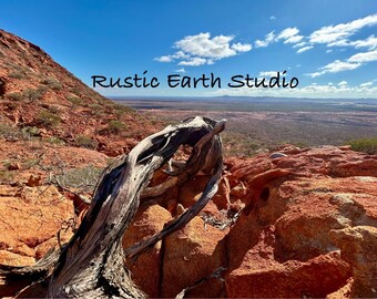 Op de afbeelding: Een landschapsfoto met een rood-bruine rotsformatie onder een blauwe lucht met wolken. Een verweerde, grijze tak ligt op de rotsen. De tekst "Rustic Earth Studio" is zichtbaar.