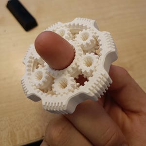 May include: A white 3D-printed gear fidget toy with multiple interlocking gears. The toy is held by a person's finger, with the finger inserted into the center of the gear mechanism. The background includes a wooden surface and a black computer mouse.