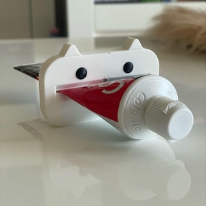 May include: A white 3D-printed cat-shaped toothpaste tube squeezer with black eyes and ears. A red and white Colgate toothpaste tube is inserted into the squeezer. The squeezer is on a white surface.