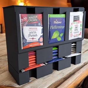 May include: A dark grey tea organiser with multiple compartments. The organiser holds tea bags in various flavours, including "Winter-Punsch" and "Zimt Apfel." The tea bags are in colourful packaging. The organiser is on a wooden table.