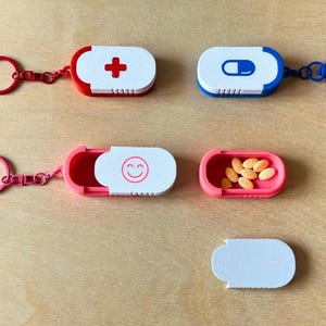 May include: A collection of colorful pill cases with keychains. The cases are red, pink, and blue, with white lids featuring a red cross, a blue pill graphic, and a smiling face. One case is open, revealing orange pills.