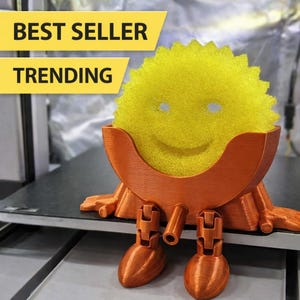May include: A whimsical orange 3D-printed sponge holder with legs and a smiling face, holding a yellow sponge. The text "BEST SELLER TRENDING" is in a yellow banner above the sponge.