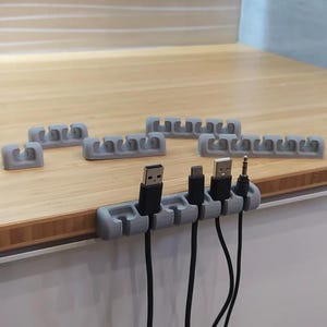 以下が含まれることがあります： 複数のスロットを備えたグレーのケーブルオーガナイザー。さまざまな黒いUSBケーブルとオーディオケーブルを保持しています。いくつかのオーガナイザーが木製の表面に配置されています。オーガナイザーはコードを管理および保護するように設計されています。