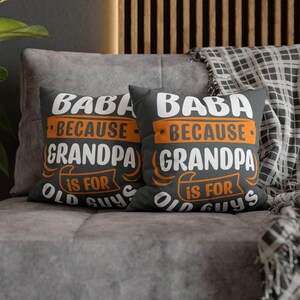 May include: Two dark gray throw pillows with the text "BABA BECAUSE GRANDPA IS FOR OLD GUYS" in white and orange. The pillows are on a gray couch with a patterned blanket.