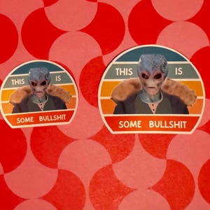May include: Two stickers featuring an alien with blue skin and a neutral expression. The stickers have the text "THIS IS" above the alien and "SOME BULLSHIT" below. The background is a red and white pattern.