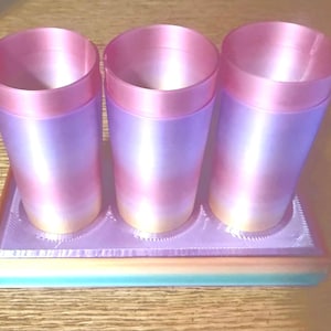 May include: Three cylindrical containers with a gradient color scheme of pink, purple, and yellow, resting on a rectangular tray. The containers have a pink rim and a smooth, glossy finish. The tray has a multi-colored base.