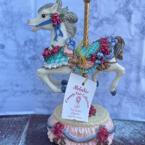 May include: A carousel horse figurine from the Melodies Fair Collection. The white horse has a gray mane and is adorned with pink and blue floral accents. The base is pink with blue ribbons and red roses. The tag reads "Press bottom of figurine to activate music."