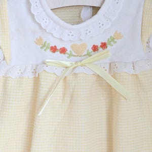 Puede incluir: Vestido de beb&eacute; con corpi&ntilde;o bordado blanco y falda de cuadros amarillos. El corpi&ntilde;o presenta un dise&ntilde;o de bordado floral y de coraz&oacute;n con un lazo de cinta amarilla. El vestido tiene un cuello con volantes y est&aacute; colgado de una percha de madera.
