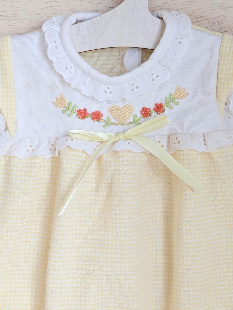Puede incluir: Vestido de beb&eacute; con corpi&ntilde;o bordado blanco y falda de cuadros amarillos. El corpi&ntilde;o presenta un dise&ntilde;o de bordado floral y de coraz&oacute;n con un lazo de cinta amarilla. El vestido tiene un cuello con volantes y est&aacute; colgado de una percha de madera.