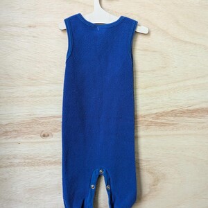 May include: A sleeveless, blue fleece baby jumpsuit with snap closures at the legs, hanging on a wooden hanger. The jumpsuit has a round neckline and a soft, textured appearance. The background is a light wood grain.