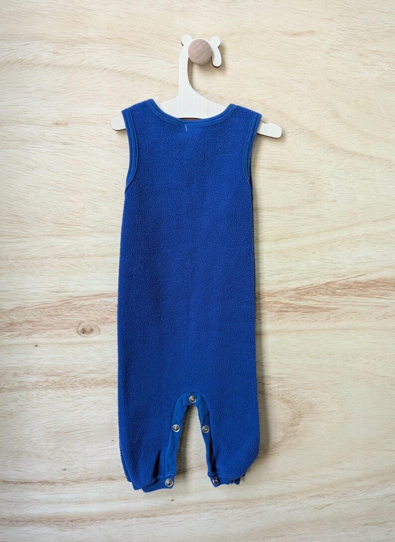 May include: A sleeveless, blue fleece baby jumpsuit with snap closures at the legs, hanging on a wooden hanger. The jumpsuit has a round neckline and a soft, textured appearance. The background is a light wood grain.