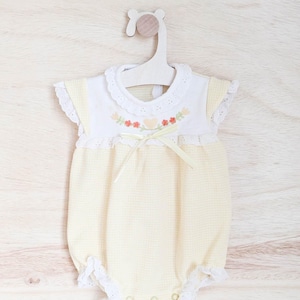 Puede incluir: Un body de beb&eacute; a cuadros amarillos y blancos con cuello blanco y ribete de encaje. El body presenta bordados florales y un lazo de cinta amarilla. Cuelga de una percha de madera sobre un fondo de madera clara.