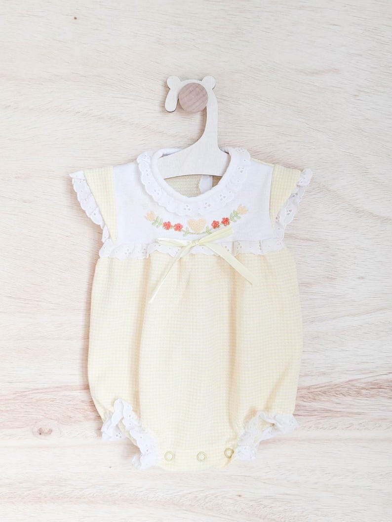 Puede incluir: Un body de beb&eacute; a cuadros amarillos y blancos con cuello blanco y ribete de encaje. El body presenta bordados florales y un lazo de cinta amarilla. Cuelga de una percha de madera sobre un fondo de madera clara.