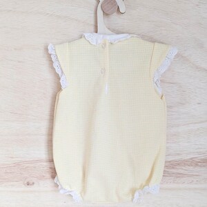 Puede incluir: Un body de beb&eacute; de cuadros amarillos con ribete de encaje blanco en las mangas y las aberturas de las piernas. El body tiene un cuello de encaje blanco y se abrocha por la espalda. Cuelga de una percha de madera.