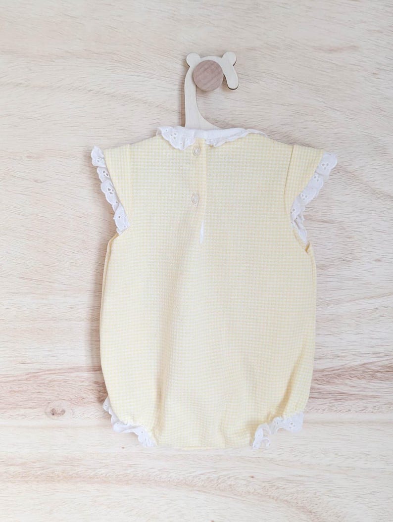 Puede incluir: Un body de beb&eacute; de cuadros amarillos con ribete de encaje blanco en las mangas y las aberturas de las piernas. El body tiene un cuello de encaje blanco y se abrocha por la espalda. Cuelga de una percha de madera.
