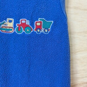 May include: Blue fleece baby sleeper with embroidered construction vehicles: a bulldozer, tractor, and dump truck. The sleeper has a round neckline and the Carter's brand label.