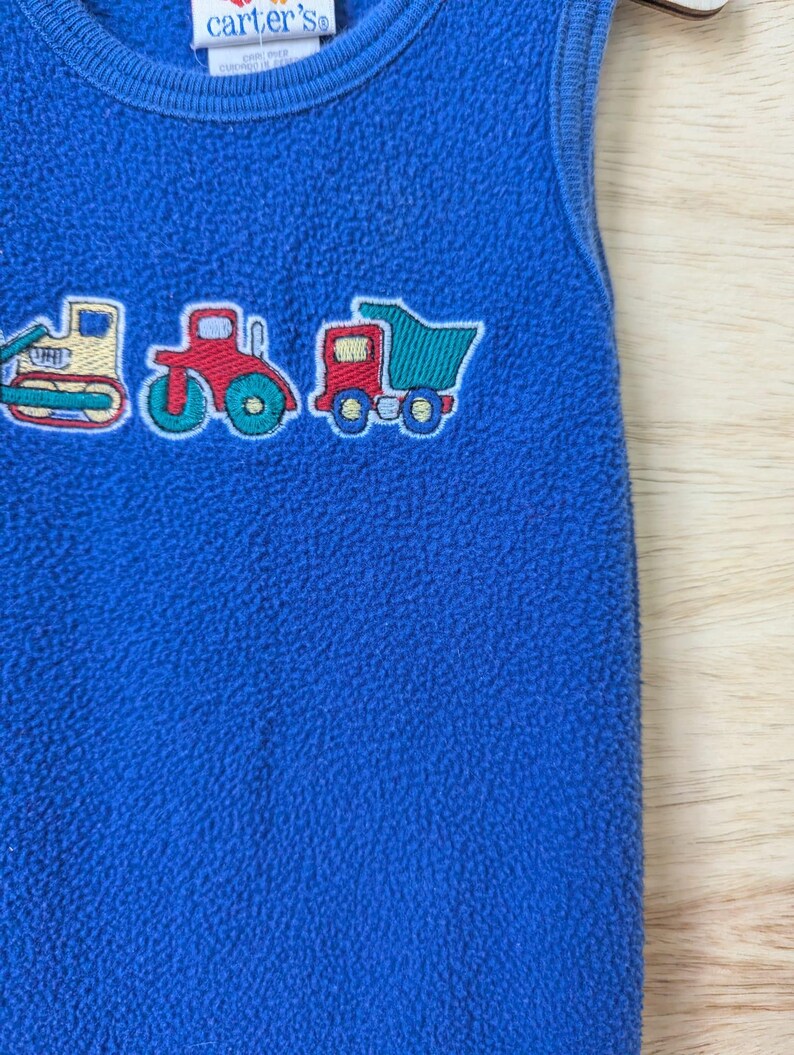 May include: Blue fleece baby sleeper with embroidered construction vehicles: a bulldozer, tractor, and dump truck. The sleeper has a round neckline and the Carter's brand label.