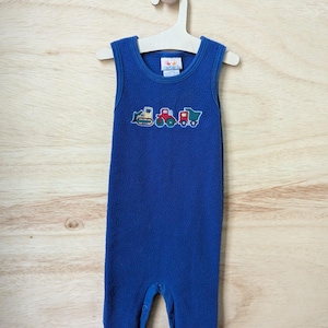 May include: A blue fleece baby romper with embroidered construction vehicles. The sleeveless one-piece garment is hanging on a wooden hanger against a light wood background. The romper features a bulldozer, a dump truck, and a cement mixer.