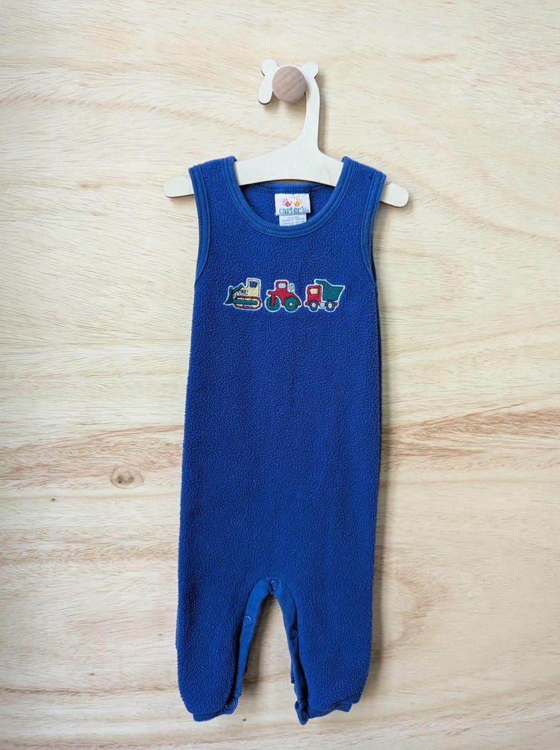 May include: A blue fleece baby romper with embroidered construction vehicles. The sleeveless one-piece garment is hanging on a wooden hanger against a light wood background. The romper features a bulldozer, a dump truck, and a cement mixer.