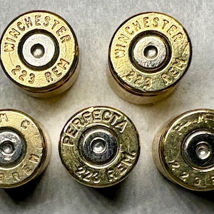 May include: Close-up of five brass bullet casings. The casings have a gold finish with silver centers. The text "WINCHESTER 223 REM" and "PERFECTA 223 REM" are visible on the casings.