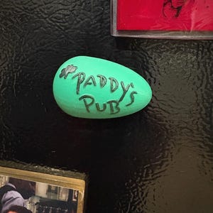 May include: A green, oval-shaped magnet with the words "PADDY'S PUB" in black lettering. The magnet is on a black surface, with other items visible in the background.
