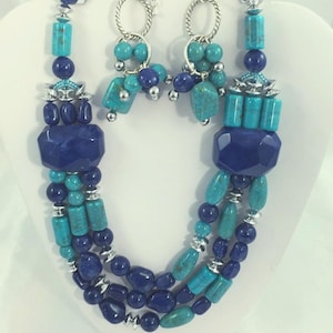May include: A three-strand necklace and matching earrings set. The necklace features large, faceted, dark blue stones, turquoise-colored beads, and silver accents. The earrings have a similar design with turquoise and dark blue beads suspended from silver hoops.