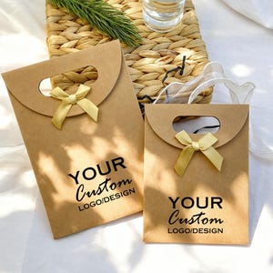 May include: Two brown paper gift bags with a curved top and a handle cutout. Each bag has a gold ribbon bow and the text "YOUR Custom LOGO/DESIGN" printed in black. The bags are on a white surface.