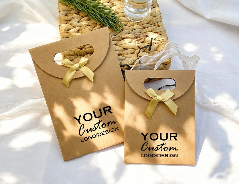 May include: Two brown paper gift bags with a curved top and a handle cutout. Each bag has a gold ribbon bow and the text "YOUR Custom LOGO/DESIGN" printed in black. The bags are on a white surface.